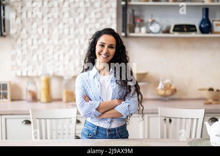 Portrait d'une femme au foyer latine heureuse posant avec les bras pliés dans la cuisine, souriant et regardant l'appareil photo, espace libre Banque D'Images