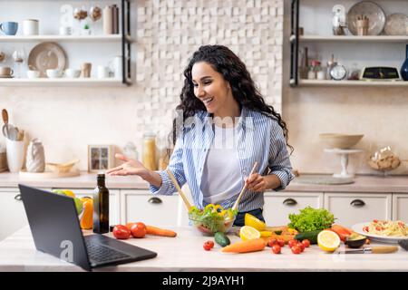 Concept de communication en ligne. Bonne femme latine préparant une salade et parlant sur chat vidéo via ordinateur portable dans la cuisine Banque D'Images