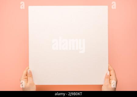 Prise de vue en hauteur de mains de femme tenant une feuille de papier vierge carrée sur une table rose. Femme mains avec maquette feuille de papier vide sur fond pastel. Les mains se sont hsocial Banque D'Images