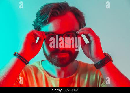 Concept de lunettes. Beau homme caucasien barbu touchant ses lunettes noires avec les deux mains et regardant la caméra. Studio gros plan portrait avec lumière contrastée - rouge et bleu. Photo de haute qualité Banque D'Images