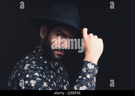 Barbu latino-américain beau homme personne touchant chapeau noir regardant sévèrement dans l'espace de copie de l'appareil photo fond noir studio tourné de gros plan moyen. Photo de haute qualité Banque D'Images