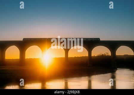 Un train Northern Rail traversant le pont ferroviaire Arthington Viaduct au lever du soleil avec la rivière Wharfe en dessous. Le soleil se lève entre les arches. Banque D'Images