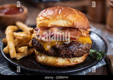 Gros plan d'un cheeseburger au bacon sur un petit pain grillé et de frites sur une assiette noire Banque D'Images