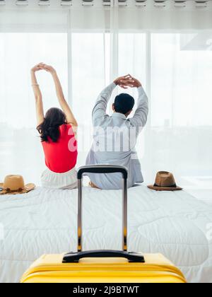 Couple de touristes, de joyeuses vacances. Vacances d'été. Vue arrière de jeune asiatique homme et femme s'étirant avec détente sur le lit avec valise près du rideau Banque D'Images