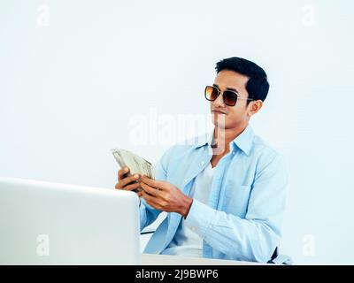 Portrait d'un jeune homme d'affaires asiatique riche et heureux en chemise bleue décontractée et portant des lunettes de soleil tout en tenant et en comptant des dollars billet d'argent Banque D'Images