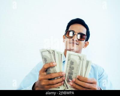 Portrait d'un jeune homme d'affaires asiatique riche et heureux en chemise bleue décontractée et portant des lunettes de soleil tout en tenant et en comptant des dollars billet d'argent Banque D'Images