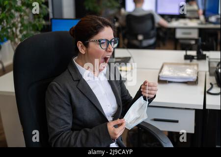 Une femme en costume d'affaires s'assoit sur un lieu de travail dans le bureau et hurle tout en prenant son masque de protection. Banque D'Images
