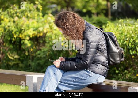 Fille aux cheveux bouclés portant un Jean et une veste imperméable en nylon assise avec un smartphone sur la banquette au parc printanier Banque D'Images