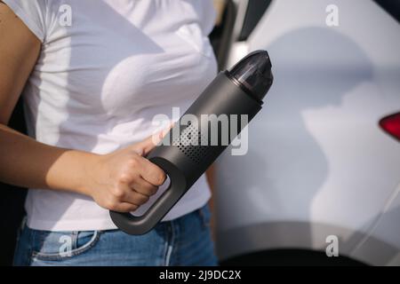 Femme tenir l'aspirateur portable devant la voiture. Belle jeune femme se prépare pour le nettoyage dans le garage. Gros plan des mains Banque D'Images