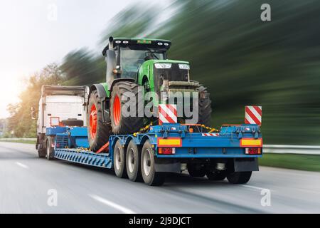 La plate-forme à plateau semi-remorque de camion industriel POV lourd transporte une grande machine de tracteur agricole moderne sur une route commune à ciel ouvert Banque D'Images