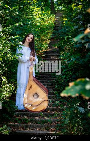 Une fille ukrainienne vêque de blanc se dresse sur une échelle en pierre au milieu de la forêt. Femme ukrainienne avec des tresses lâches avec un instrument de musique bandura Banque D'Images