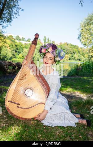 Jeune femme ukrainienne assise près de la rivière en robe nationale et portant un instrument de bandura à cordes nationales ukrainiennes. Banque D'Images