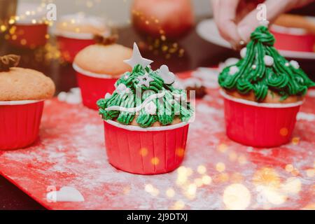 Petits gâteaux en forme d'arbre de Noël avec glaçage à la crème sur les lumières de noël et le fond de fête. Homme mains cuisine de Noël cupcakes traitement pour fami Banque D'Images