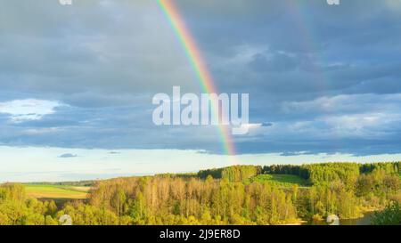 Double arc-en-ciel dans un ciel bleu ciel nuageux après la pluie au coucher du soleil. Un véritable arc-en-ciel lumineux contre un paysage forestier vallonné au début du printemps. Concept de précipitation Banque D'Images