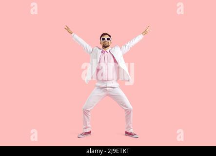 Photo pleine longueur d'une jeune fashionista dansant dans le studio. Joyeux jeune homme beau dans un costume blanc, une chemise rose, des baskets roses et des lunettes de soleil Banque D'Images