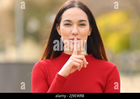 Vue de face d'une femme heureuse en rouge demandant le silence avec le doigt sur les lèvres dans la rue Banque D'Images