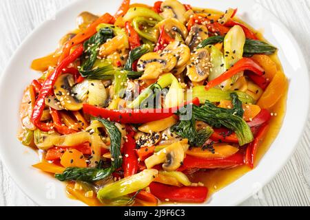 Gros plan de la sauce de légumes frite de chou de bok choy, de poivrons rouges, de champignons, d'oignons, de carottes et de sauce sur une assiette blanche sur une table en bois blanc Banque D'Images