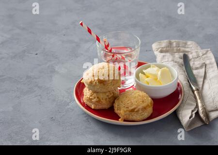 Scones maison fraîchement cuits avec du fromage et des herbes sur une assiette rouge avec du beurre sur un fond texturé gris. Petits pains traditionnels anglais Banque D'Images