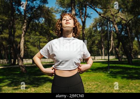 Belle jeune femme décontractée portant un t-shirt blanc et un pantalon de yoga noir appréciant la nature respirant l'air frais méditant sur le parc de la ville, à l'extérieur. Banque D'Images