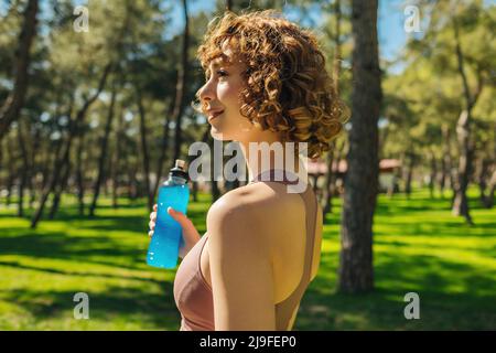 Vue latérale de la femme sportive à tête rouge portant un soutien-gorge de sport et tenant une boisson énergétique ou énergétique tout en se tenant sur le parc vert de la ville, à l'extérieur. En-cas sains, Banque D'Images