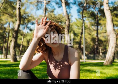 Jogging sport fatigué épuisé fille élimine la transpiration du front. Assis sur l'herbe sur le parc vert de la ville, à l'extérieur. Concept de vie saine et de sports de plein air Banque D'Images