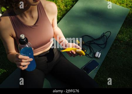 Vue rognée d'une femme sportive à tête rouge portant un soutien-gorge de sport et contenant une bouteille de boisson énergétique ou de puissance et une banane. Des aliments sains, des collations et des services d'après-vente en extérieur Banque D'Images