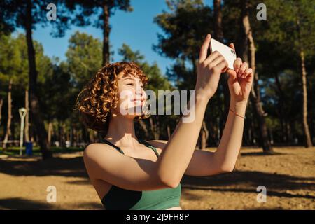 Jeune femme à tête rouge portant des vêtements de sport prenant des photos avec un smartphone sur le parc de la ville, à l'extérieur. Banque D'Images