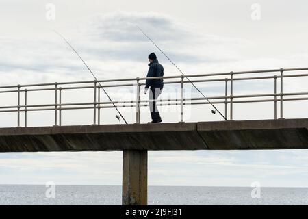 Un homme se tient sur un quai entre deux cannes à pêche dans le comté de Northumberland, au Royaume-Uni. Banque D'Images