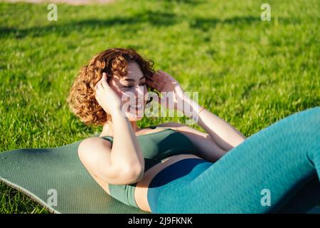 Jeune heureuse redhead femme portant des vêtements de sport faire des sit-ups abs craquements exercice à l'intérieur allongé sur le tapis de yoga. Entraînement sportif de routine, physi matin Banque D'Images