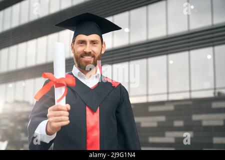 Vue de face d'un étudiant diplômé de l'école secondaire. Jeune homme attrayant portant un mortier et une robe de troisième cycle, debout, montrant le diplôme, souriant. Concept de réussite. Banque D'Images