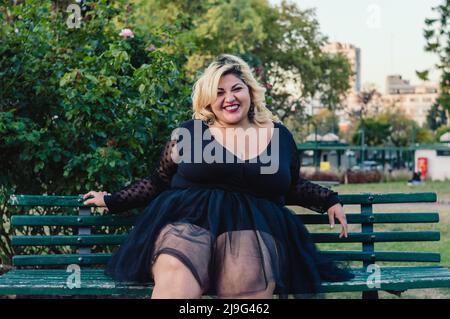 belle jeune femme de taille plus avec robe de tulle noire, assise sur un banc en bois dans le parc heureux de regarder l'appareil photo et rire. Banque D'Images