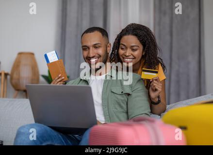 Un heureux couple afro-américain utilisant un ordinateur portable et réserver un hôtel avec une carte de crédit, assis avec des valises, prêt à partir Banque D'Images