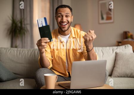 Jeune homme excité avec passeport et billet gestante OUI, en utilisant l'ordinateur portable pour acheter la visite chaude à l'agence de voyage web de la maison Banque D'Images