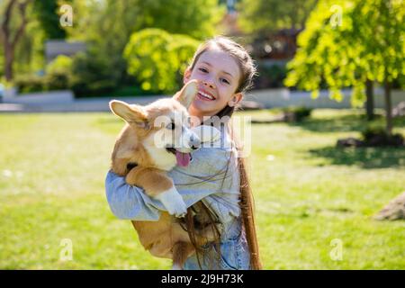 Fille tenant son chiot corgi dans ses bras. Une fille joue avec son chiot corgi sur une pelouse verte par temps ensoleillé. Banque D'Images