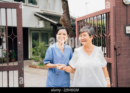 Infirmière féminine optimiste en uniforme bleu soutenant une femme asiatique âgée tout en marchant ensemble à travers la porte à l'extérieur de la maison Banque D'Images