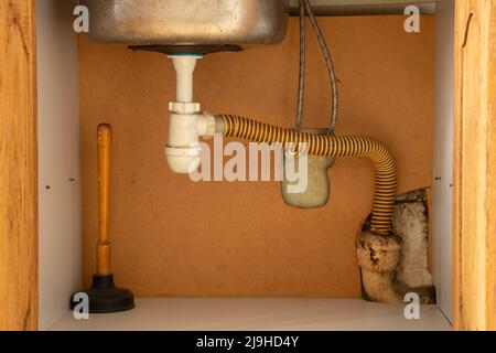Un piston se tient dans un placard sous l'évier dans la cuisine, nettoyant les tuyaux bouchés, un ancien nettoyeur de tuyaux Banque D'Images
