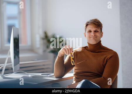 Homme d'affaires souriant tenant des lunettes assis au bureau Banque D'Images