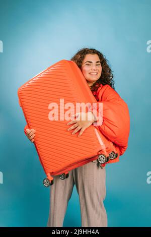 Bonne jeune femme portant une valise orange sur fond bleu Banque D'Images