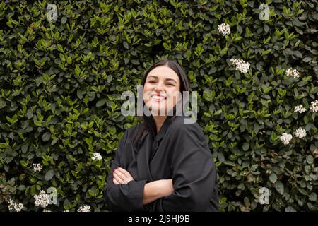 Une femme heureuse portant un blazer noir debout devant le mur de plantes vertes Banque D'Images