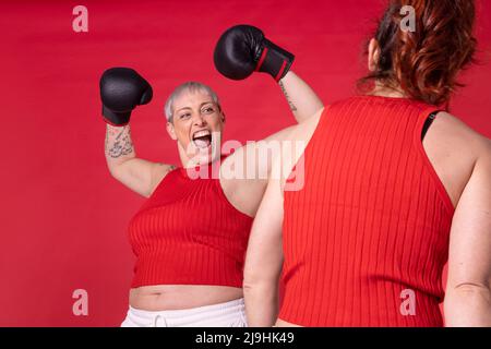 Femme portant des gants de boxe fléchissant les muscles regardant l'ami contre fond rouge Banque D'Images