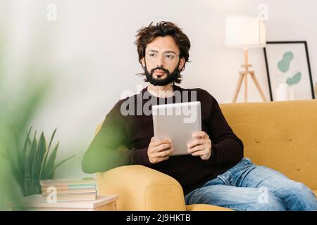 Homme barbu attentionné avec tablette PC assis sur un canapé dans le salon à la maison Banque D'Images