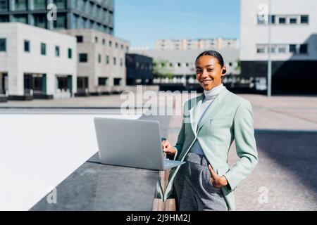 Femme d'affaires souriante avec ordinateur portable dans le quartier du centre-ville par beau temps Banque D'Images