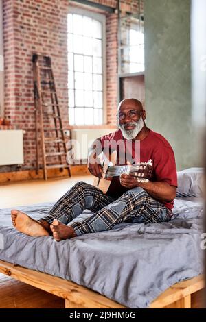 Homme mature avec lunettes jouant de la guitare sur le lit à la maison Banque D'Images