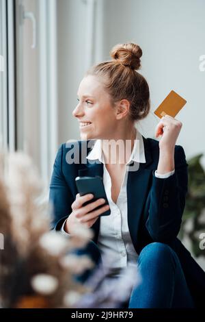 Femme d'affaires envisageant de tenir un smartphone et une carte de crédit au bureau Banque D'Images