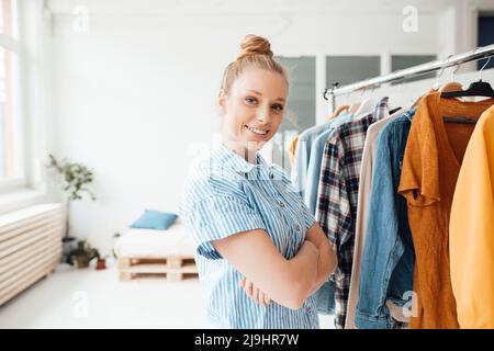 Styliste souriant avec bras croisés debout près du porte-vêtements du studio Banque D'Images