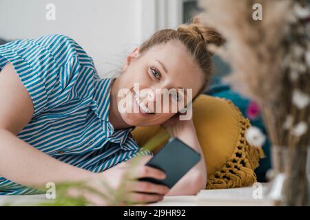 Jeune freelance souriante avec un smartphone allongé sur le lit à la maison Banque D'Images