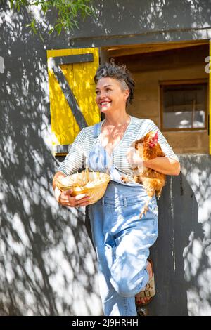 Fermier mûr souriant tenant la poule et le bol d'oeuf pendu sur le coop de poulet Banque D'Images