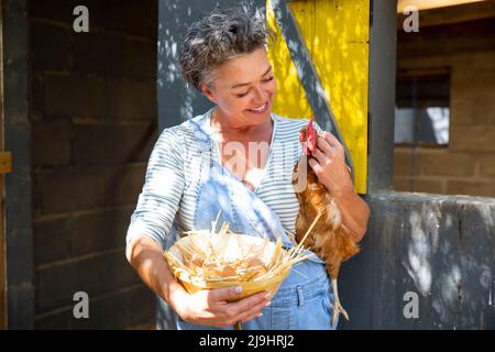 Propriétaire de ferme mûr souriant transportant la poule et le bol d'œufs devant le coop de poulet Banque D'Images