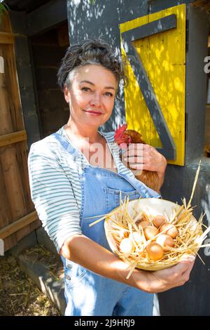 Femme mûre souriante avec une poule et un bol d'œufs debout devant le coop de poulet Banque D'Images