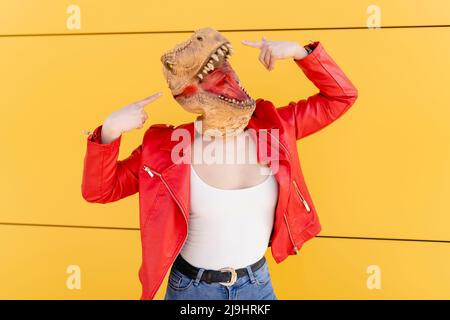 Femme joueur portant un masque de dinosaure pointant devant le mur jaune Banque D'Images